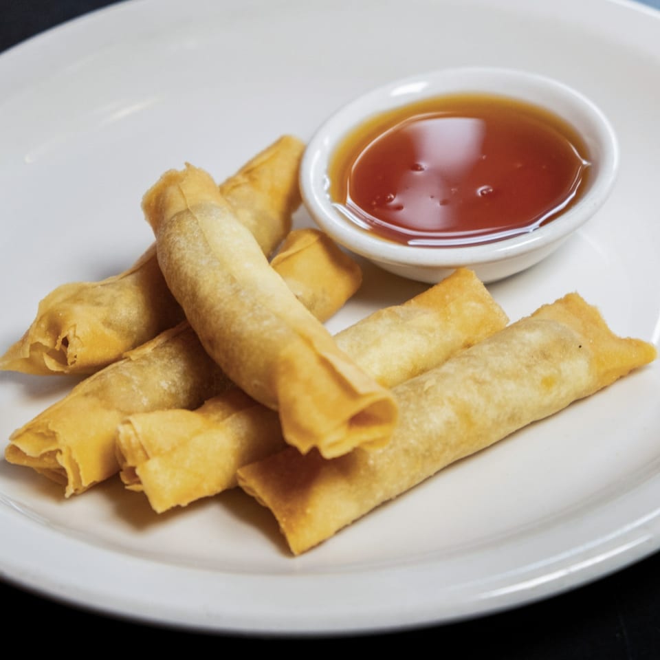 Golden-fried Thai spring rolls with a side of sweet chili dipping sauce on a white plate.