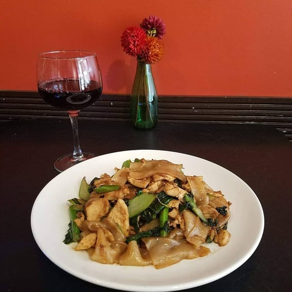 A plate of Pad See Ew with chicken and greens, a glass of red wine, and a small vase of flowers.