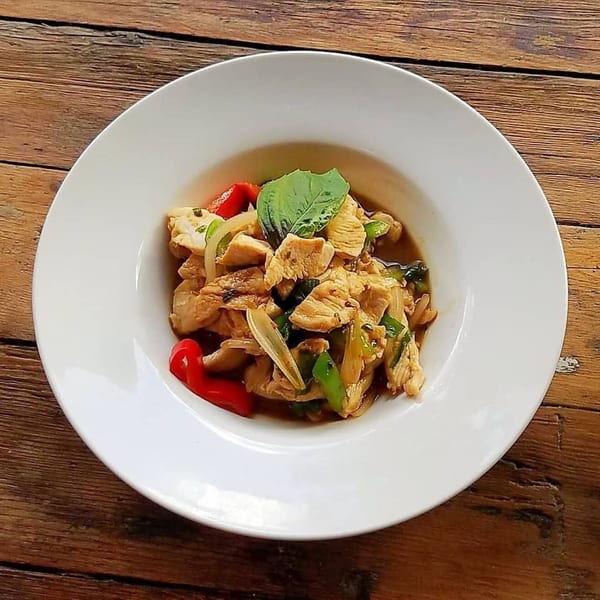 Stir-fried chicken with red bell peppers, onions, and basil in a white bowl on a wooden table.
